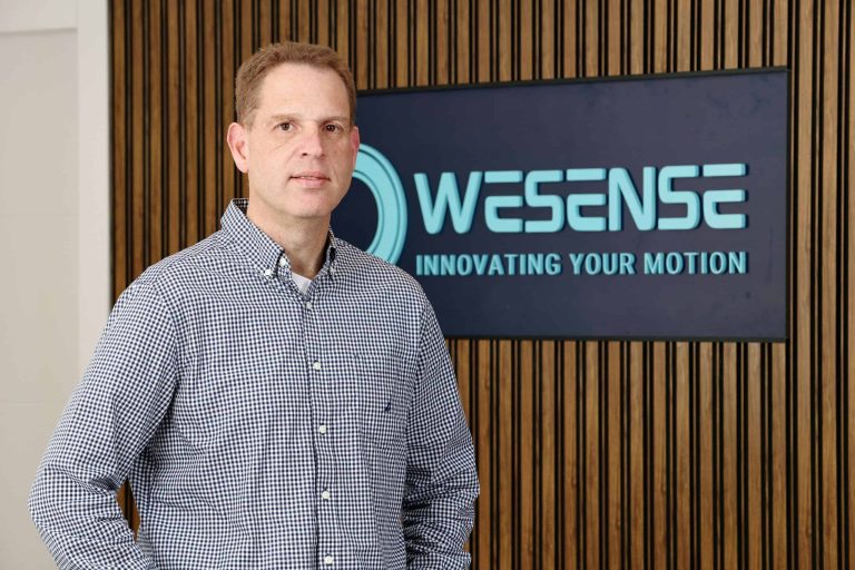 Professional man standing in front of WeSense logo on wooden wall.